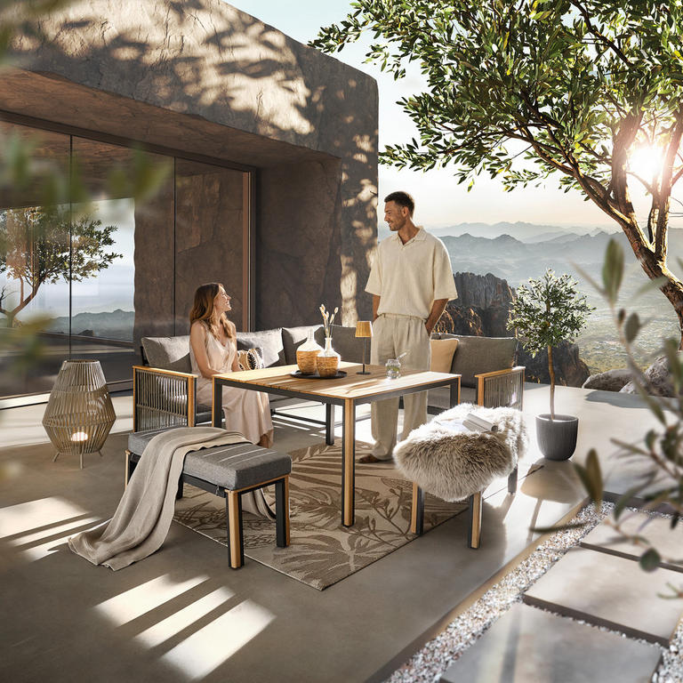 Zwei Personen sitzen und stehen an einem modernen Gartentisch auf einer Terrasse mit Blick auf Berge und Pflanzen.